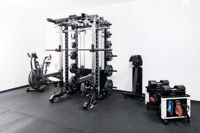 Gym equipment including a Smith machine rack attachment and weight rack on a black floor with white walls.