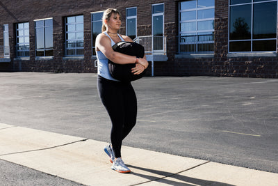 Woman performing a stone sandbag carry outdoors on a sidewalk with a building in the background