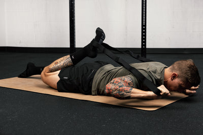 Person lying on a  yoga mat in a gym setting with visible tattoos using stretching band for quad