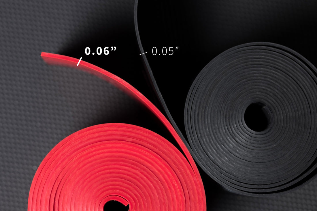 Overhead view a pair of rolled up floss bands showing that the black has a 0.05" thickness and the red has a 0.06" thickness.